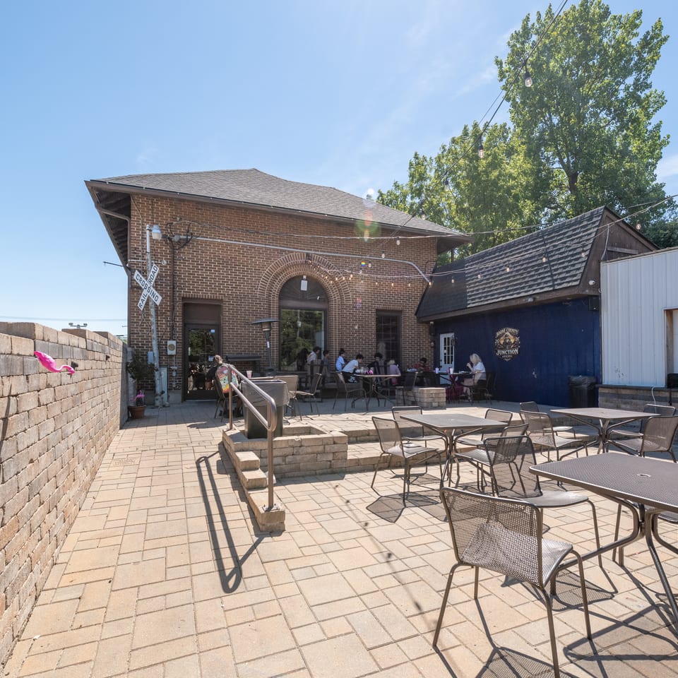 Outdoor restaurant patio with tables, chairs, string lights, brick historic building, and diners beneath a sunny blue sky.